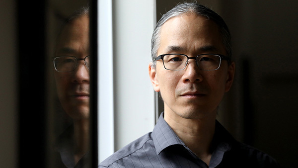 A headshot of author Ted Chiang, shadowed, with a reflection of his face on the left of the screen.